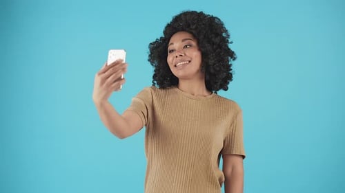 Attractive Young Woman Stands on a Blue Background and Communicates with Someone Through a Phone