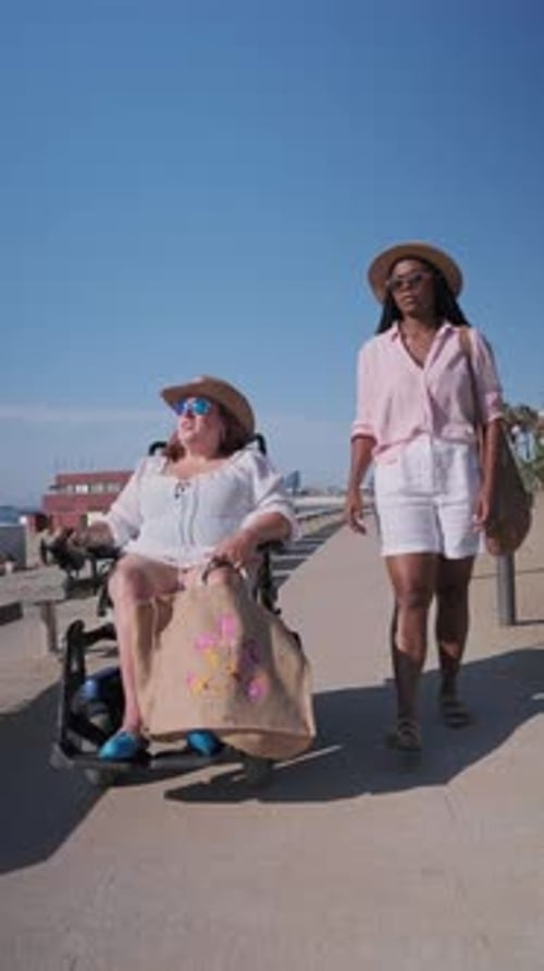 Woman Pushing Friend in Wheelchair on Beach Boardwalk in Slow Motion
