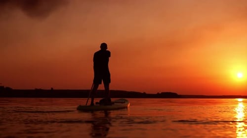 Aufnahme eines Mannes beim Supboarden am Fluss bei Sonnenuntergang. Der Sportler rudert langsam mit einem langen Paddel
.