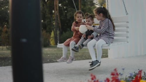 Three Little Girls Having Fun on a White Swing in a Quiet Treelined Park Background Sisters Eating