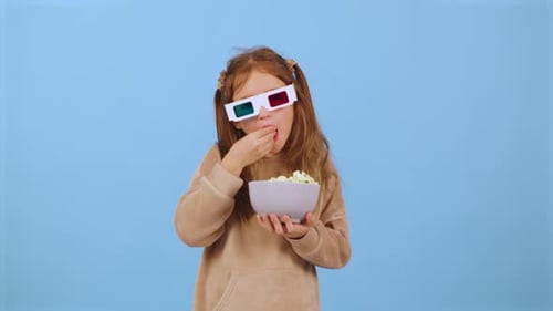 Young Girl Enjoys Popcorn While Wearing 3D Glasses in Front of a Blue Background During a Fun Movie