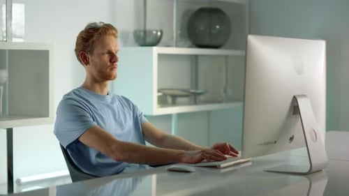 Rothaariger Mann tippt auf einem Desktop-Computer im Home-Office, Fernansicht des Arbeitsplatzes