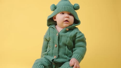 Portrait of a Cute Baby in a Warm Knitted Jumpsuit with a Hood on a Yellow Background