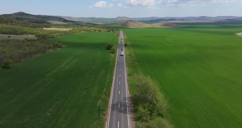 Cars driving steadily along a straight road, cutting through vibrant green agricultural fields, dron
