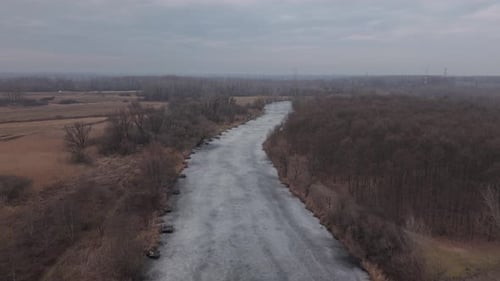 Aerial drone view of a long frozen river flowing through a quiet winter countryside. Leafless