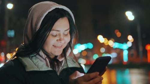 Portrait of a Young Woman Texting on Her Smartphone on a City Street at Night