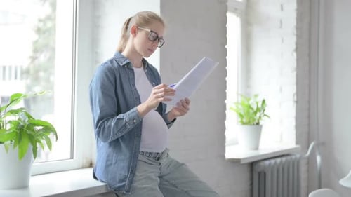 Pregnant Young Woman Reading Documents While Sitting in Window