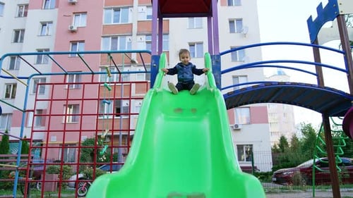 Little kid sits on the big green slide. Happy kid slides down approaching camera. Low angle view.
