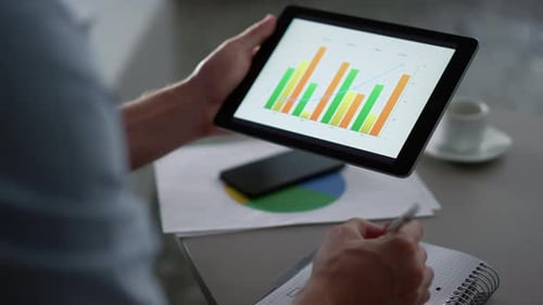 Man reviewing tablet with graphs, writing in notebook