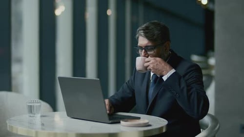 Senior Businessman Drinking Coffee as Working on Laptop at Cafe Table