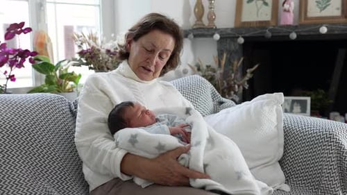 Senior Woman Holds Newborn Infant on Couch Indoors
