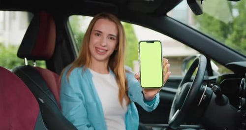 Beautiful young woman sitting in a car on drivers seat and showing on camera smartphone