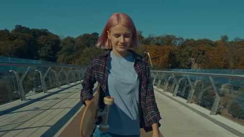Joyful Pretty Pink Haired Woman with Skateboard Walking on City Bridge at Sunrise