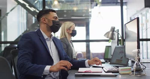 Homme d'affaires métisse portant un masque facial et utilisant un ordinateur avec un collègue travaillant en arrière-plan