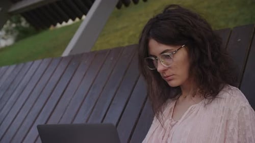 Handheld Close Up of a Curly Caucasian Woman Working on Her Laptop Computer on a Park Bench