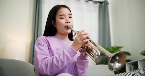 A young woman practices playing the trumpet while sitting on a sofa in a cozy living room.