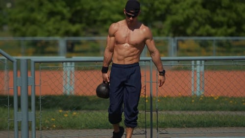 Young Handsome Man in Black Cap and Sunglasses Takes Kettlebell and Goes to Training on Outdoor