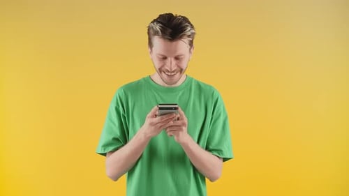 A Smiling Guy in a Green Tshirt Stands Against a Yellow Wall with a Phone in His Hands A Young Man