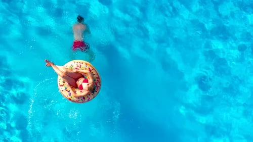 Casal se divertindo na piscina, o homem mergulha e a mulher se deita em um donut