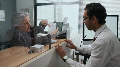Elderly Woman Arguing with Bank Employee Across Glass Teller Window