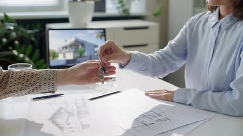 Real Estate Agent Handing Keys to Client over Table with Architectural Drawings