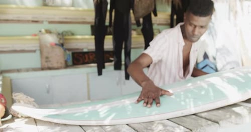 African american man preparing surfboard on the counter of surf rental beach shack, slow motion