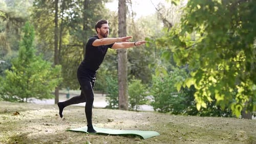 Der Mann führt eine Yoga-Balance-Pose auf einer Gymnastikmatte in einem ruhigen Park im Freien durch. Umgeben von grünen Bäumen,