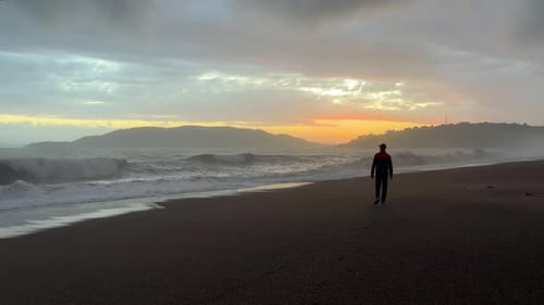 A Man Walks Along a Sandy Beach Along a Stormy Sea Against the Backdrop of a Beautiful Golden Sunset