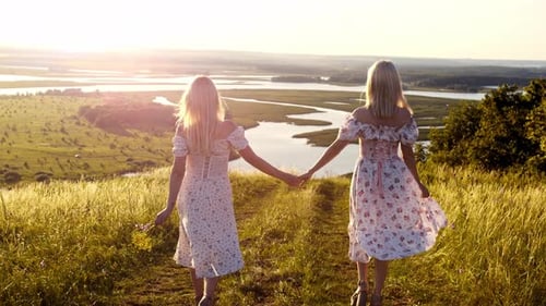 Two Blonde Young Women Hold Hands and Go Down the Hill to the River