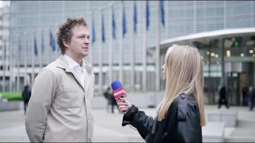 Periodista entrevistando a un hombre frente a un edificio moderno con banderas en una escena mediática