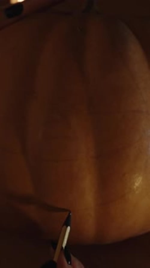 Vertical video. In the Dark with Flickering Candlelight, a Woman Draws Mouth and Eyes on a Pumpkin.