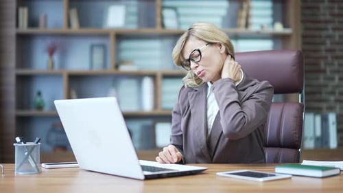 Tired mature businesswoman feeling neck pain while working on laptop at workplace at desk in office.