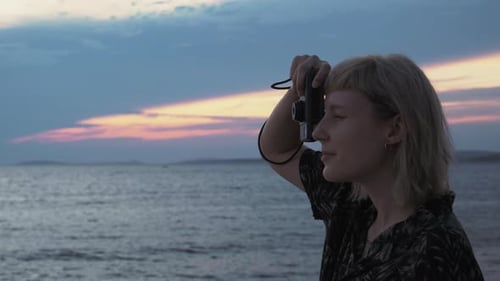 Close up shot of a young blonde woman taking photos on film of the amazing sunset near the sea.