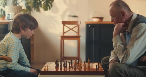 Seitliche Ansicht eines niedlichen Jungen im Teenageralter, der im hellen Wohnzimmer Schach gegen den alten Großvater spielt