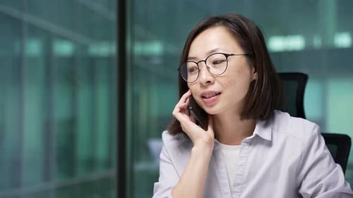 Confident asian businesswoman talking on mobile phone while sitting at workplace in office.