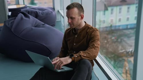Young man with laptop sitting on floor by the window. Man an is working remotely