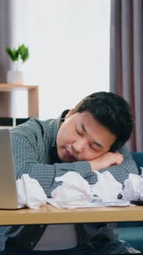 Tired Adult Asian Man Sleeping on the Office Table Covered with Many Crumpled Papers in Contemporary