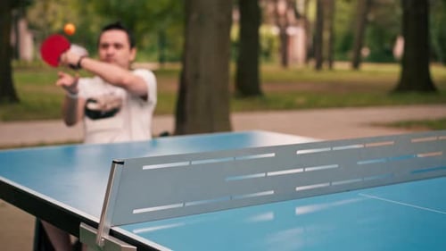 A man in a wheelchair plays table tennis in a city park practices strokes with a ping pong racket