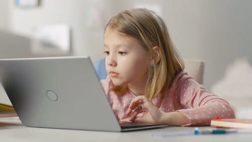Cute Young Girl Uses Laptop Computer while Sitting at the Table in Her Light Room.