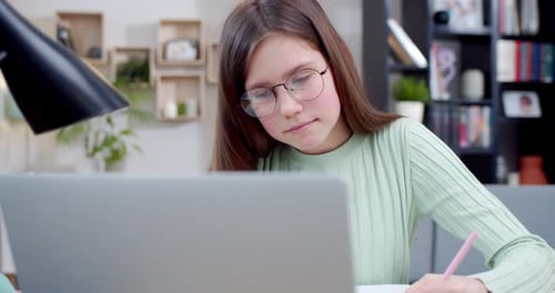 Close Up of Caucasian Teen Schoolgirl in Glasses Sitting at Laptop Writing Exercise in Copybook and