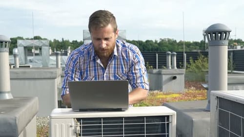 Frustrated man checks air conditioning unit on roof with laptop in the city