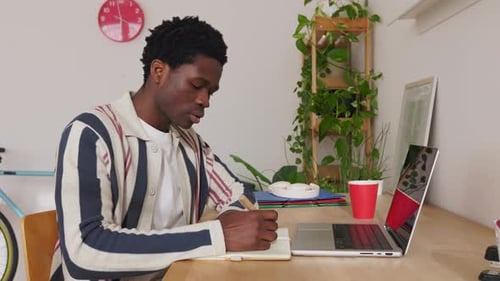 Focused Black Man Taking Notes While Working on Laptop From Home