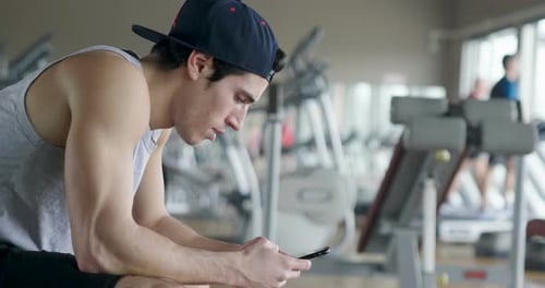 Portrait of a guy in a gym while resting between a workout and the other uses his phone to watch s