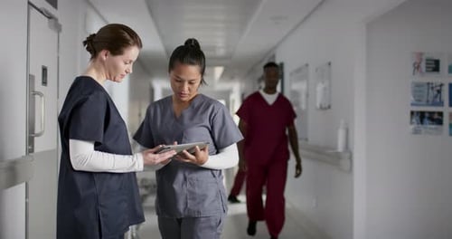 Diverse female doctors discussing work, using tablet in corridor at hospital, slow motion