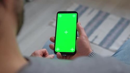 Young Man Sitting at Home Holding Smartphone Green Mockup Screen in Hand Male Person Using Chroma