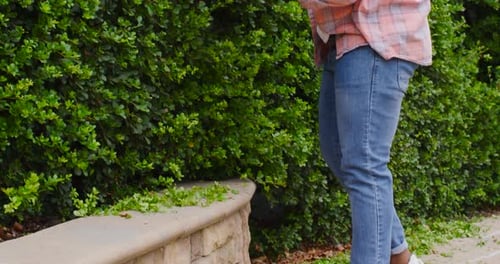 African American man starting red hedge trimmer, neatening hedge along stone bench, copy space