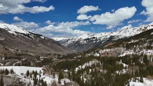 Aerial View slowly panning over pine trees with snowcapped mountains in the background with partly c