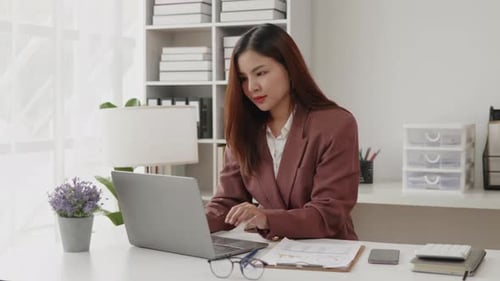 Geschäftsfrau im braunen Anzug arbeitet an einem Laptop mit glücklichem Gesicht in Büroräumen.