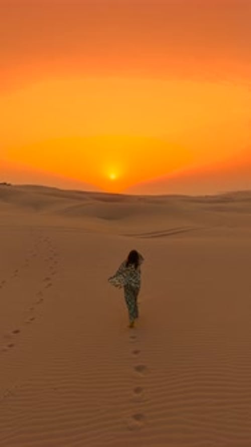Aerial View of a Young Woman Walking Across Sand Dunes Towards a Bright Sunset in the Desert of