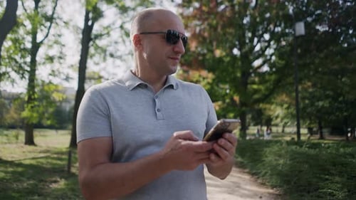 Adult Man in Park on Smartphone Wearing Sunglasses and Casual Shirt Surrounded By Nature